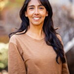 Headshot of Lucia Valdez, a medium skinned woman with long brown hair and a big smile. She is wearing a light brown long sleeved shirt and is in an outdoor setting.