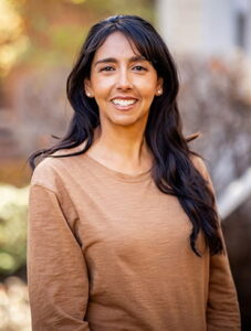 Headshot of Lucia Valdez, a medium skinned woman with long brown hair and a big smile. She is wearing a light brown long sleeved shirt and is in an outdoor setting.