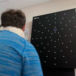Rory is shown with his back towards the viewer. He is wearing a blue shirt and a neck brace and looking at a black board on the wall as part of his stroke recovery therapy.