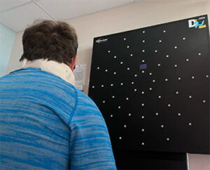 Rory is shown with his back towards the viewer. He is wearing a blue shirt and a neck brace and looking at a black board on the wall as part of his stroke recovery therapy.