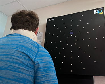 Rory is shown with his back towards the viewer. He is wearing a blue shirt and a neck brace and looking at a black board on the wall as part of his stroke recovery therapy.