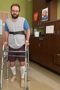 Rory Polera is pictured in a hospital room. He is walking with the assistance of a walker. He is a light skinned male wearing a grey shirt with blue sleeves, brown shorts, and tall white socks. 