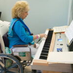 A light skinned woman with blond hair and a blue shirt playing the piano while seated in a wheelchair.